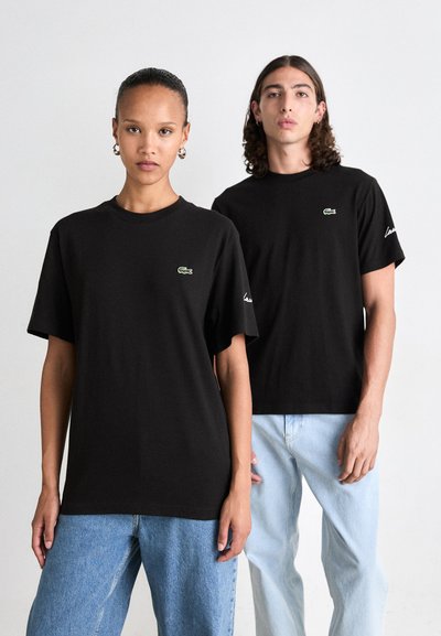 Два модела, облечени в черни тениски Lacoste и светло сини дънки, стоящи на неутрален бял фон.