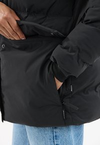 Hånd i lommen på en sort puffjakke, den anden hånd løfter jakke-flappen, person iført blå jeans.