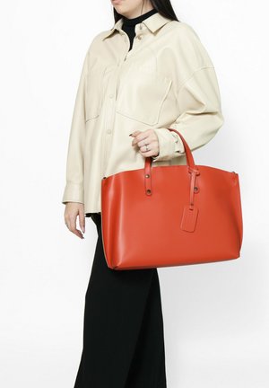 Borsa tote in pelle rossa con doppio manico, forma rettangolare, arricchita da un'etichetta da viaggio e dettagli in metallo.
