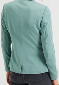Miętowo zielona dopasowana marynarka o gładkiej fakturze, z pojedynczym rozcięciem z tyłu i dopasowanymi szwami, uzupełniona długimi rękawami.