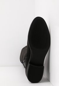 Botín negro de ante con suela de goma texturizada y surcos horizontales, detalle de cremallera lateral y sutilezas en la costura.