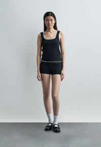 Jeune femme debout, portant un débardeur noir et un short bordé de blanc, des chaussettes blanches et des chaussures Mary Jane noires, sur un fond uni.