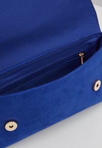 Blå tygväska med en slät textur, utrustad med ett blixtlåsförsett fack och detaljer i guldfärgad metall. Insidan är fodrad med matchande blått material.