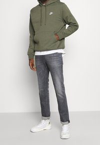 Olivgrön Nike-hoodie med framficka och dragsko, kombinerad med grå jeans och vita sneakers. Enkel design; slät textur.