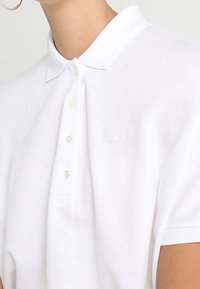 Polo blanco hecho de tejido texturizado, con un cuello clásico, mangas cortas y un pequeño logo bordado en la parte superior izquierda.