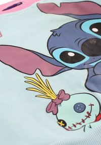 Sweat-shirt bleu clair avec un graphique d'un personnage bleu aux grandes oreilles et une poupée verte cousue avec un nœud rose. Texture de tissu doux.