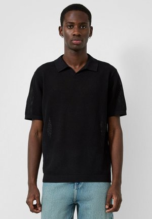 Junger Mann mit kurzen Haaren und Ohrringen, trägt ein schwarzes kurzärmeliges Strick-Poloshirt und hellblaue Jeans, steht vor einem einfarbigen Hintergrund.