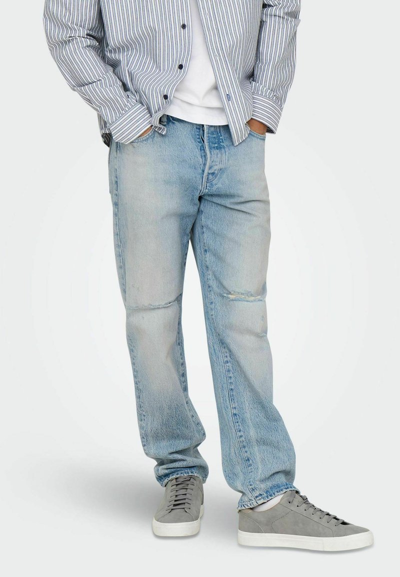 Jean en denim bleu clair à coupe décontractée, avec des sections décolorées et un léger effet usé, associés à des baskets grises et une chemise rayée.