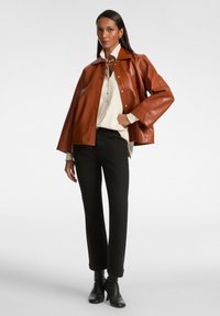Chaqueta de cuero sintético marrón con un amplio cuello y botones a presión, combinada con una camisa clara, pantalones negros y botines de tacón negros.