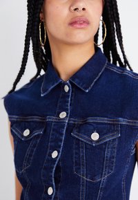 Chemise sans manches en denim, bleu foncé avec un col pointu, dotée de deux poches avant et de boutons argentés avec des coutures jaunes contrastées.
