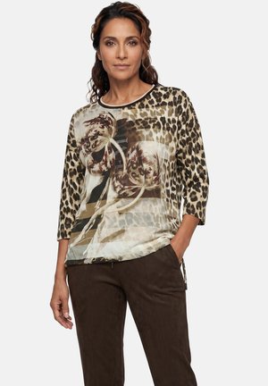 Donna che indossa una maglia con stampa leopardata e motivo floreale con maniche a tre quarti, abbinata a pantaloni marrone scuro, in piedi con una mano in tasca.