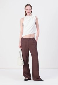 Biely top bez rukávov s detailom uzla, skombinovaný s hnedými širokými nohavicami. Modelka drží malú bielu kabelku a má na sebe čierne špicaté topánky.