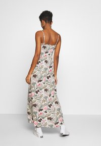 Robe longue à fines bretelles, avec un motif floral vert, rose et marron sur un fond clair. Tissu fluide et longueur sous le genou.