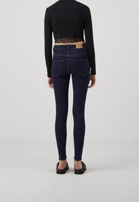 Mujer vestida con jeans ajustados de color azul oscuro y una blusa negra de manga larga y recortada con detalles de encaje, de pie contra un fondo claro y liso.