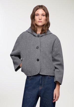 Giacca cropped in misto lana grigia con cappuccio, caratterizzata da tre bottoni neri, maniche ampie e orlo arrotondato. Abbinata a pantaloni in denim blu.