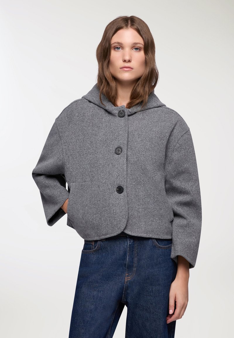 Giacca cropped in misto lana grigia con cappuccio, caratterizzata da tre bottoni neri, maniche ampie e orlo arrotondato. Abbinata a pantaloni in denim blu.