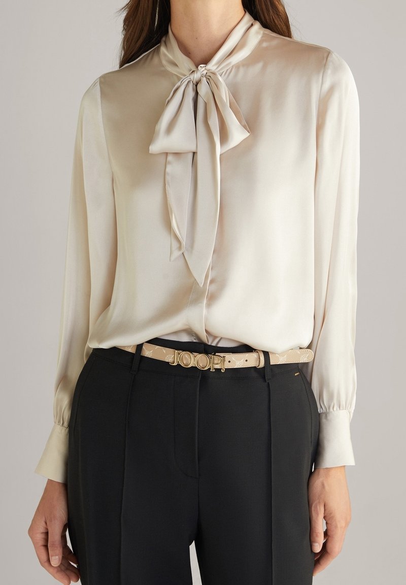 Blusa di seta in bianco sporco con dettaglio del nodo al collo, maniche lunghe con polsini abbottonati, abbinata a pantaloni neri su misura e una cintura beige con logo.