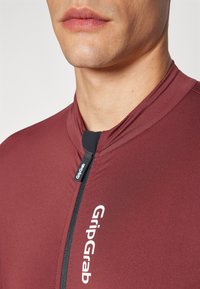 Burgunderfarget sykkeldrakt med høy krage, halv glidelås foran og GripGrab-logo. Jevn stoff med en tettsittende design og minimal sømdetaljering.