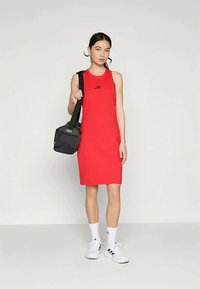 Rdeča brezrokavnika obleka z logotipom Adidas, kombinirana z majhno črno torbico čez ramo in belimi supergami. Ozadje je v nevtralni barvi.