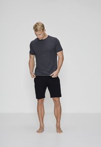 Blond mand iført mørkegrå t-shirt og sorte shorts står barfodet på hvidt gulv mod en ensfarvet lys grå baggrund og kigger ned.