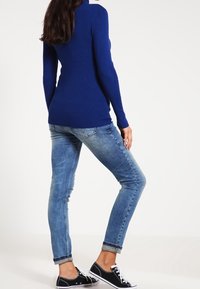 Suéter de punto acanalado azul real, diseño ajustado. Vaqueros desgastados de color azul claro, corte ajustado con bajos doblados. Zapatillas de lona negras.