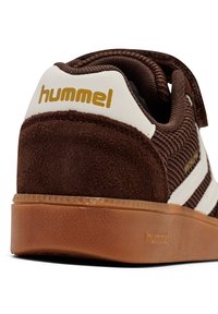 Brązowy zamszowy tenisówka z białymi akcentami, z teksturowanym korpusem, złotym logo i gumową podeszwą. Zapięcie na rzep.