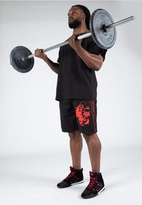 Schwarzes T-Shirt mit kurzen Ärmeln und schwarze Shorts mit roten Grafiken. Schwarze Sportschuhe mit roten Akzenten. Hält eine Langhantel mitGewichten an den Enden.