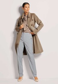 Hellbrauner doppelt geknöpfter Mantel, beige Bluse mit Schleife und hellgraue maßgeschneiderte Hose. Model trägt hautfarbene High Heels und posiert vor einem schlichten Hintergrund.
