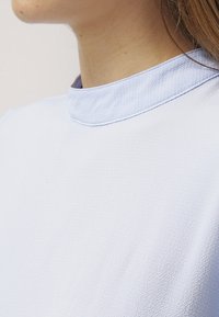 Top azzurro chiaro con superficie texturizzata, dotato di un colletto piccolo e di una finitura liscia e leggermente lucida. Ingrandimento della scollatura.