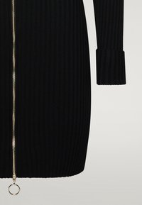 Musta ribbineulottu mekko, jossa kokopitkä vetoketju ja kullanvärinen vetoketju sekä vartalonmyötäinen siluetti. Pitkät hihat, joissa ribbineulotut mansetit.