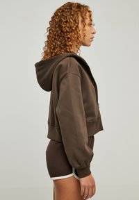 Brun cropped hettegenser laget av myk stoff, med frontglidelås og stor hette. Modellen har matchet den med brune shorts.