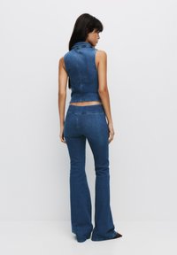 Denimoutfit bestaande uit een mouwloze top met een hoge kraag en flared jeans, beide in medium blauw, met zichtbare stiksels en naden.