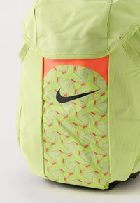 Sección de mochila verde claro con un patrón geométrico en neón, un pequeño panel naranja y un logo negro de Nike en el bolsillo frontal.