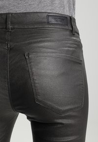 Pantalones vaqueros de mezclilla negra con un acabado brillante, que cuentan con un bolsillo trasero y una etiqueta de cuero con la marca "VERO MODA". Textura suave.