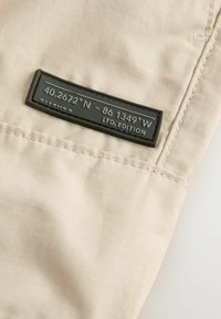 Beżowa tkanina o gładkiej fakturze z czarną etykietą, na której widnieją współrzędne oraz napis "LTD. EDITION." Widoczne są również szwy.