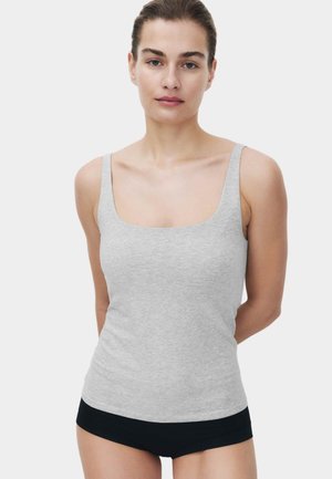 Kvinde med brunt hår bundet tilbage iført en lysegrå tank top og sorte shorts, stående med armene bag ryggen mod en ensfarvet baggrund.