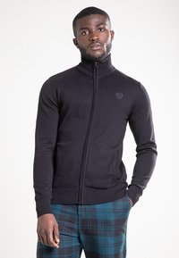 Zwarte trui met rits en een hoge kraag, geribbelde manchetten en zoom, met een klein logo op de borst. Gladde textuur, aansluitend ontwerp.