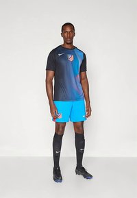 Mies, joka seisoo musta ja sininen jalkapallopaita, siniset shortsit, mustat polvimittaiset sukat ja mustat nappula-kengät yksinkertaista taustaa vastaan.