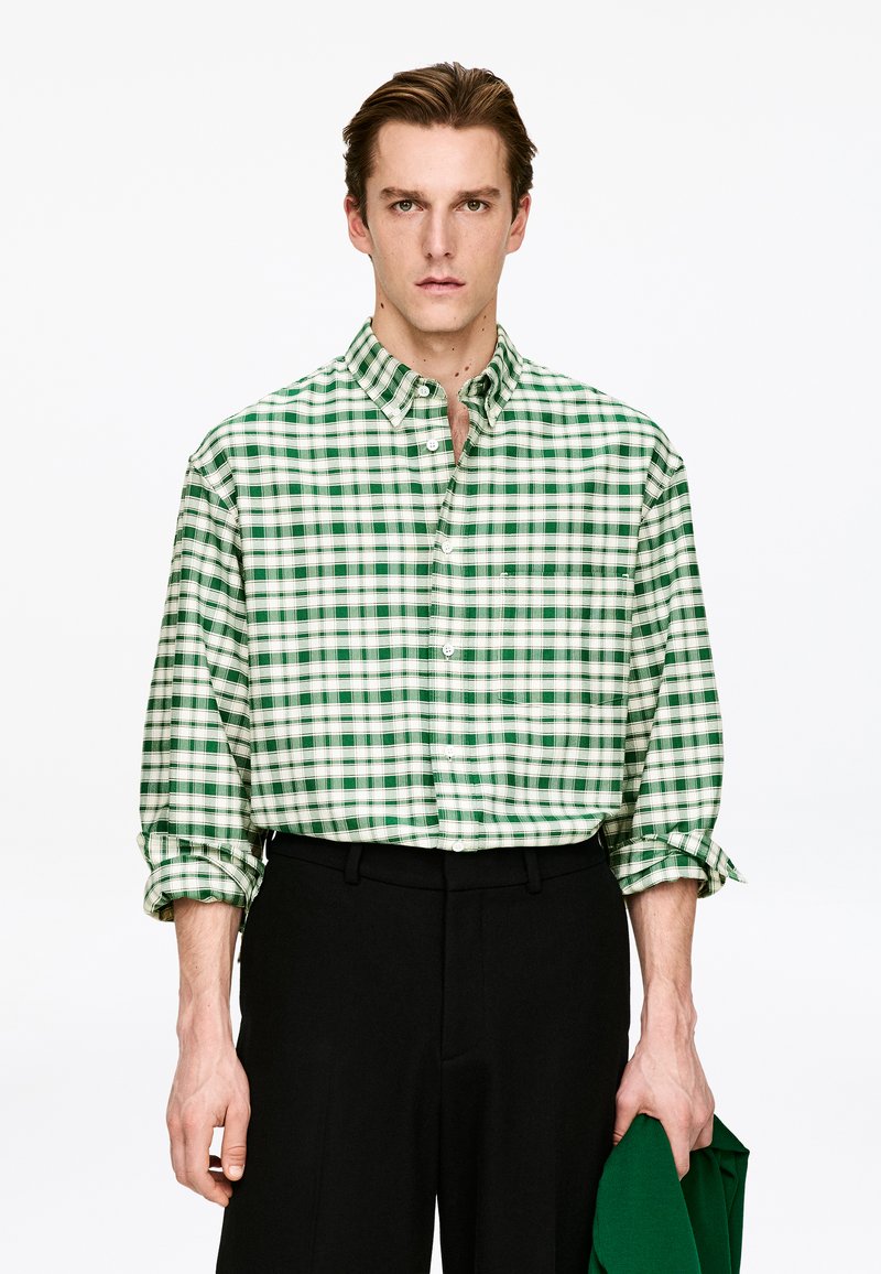 Chemise à carreaux verts et blancs en tissu léger, avec col boutonné et manches retroussées, associée à un pantalon noir.