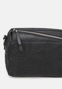 Borsa in pelle nera con motivi circolari in rilievo, chiusura a zip e forma piatta e rettangolare. Superficie testurizzata con hardware in argento.
