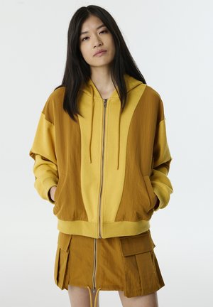 Le sweat à capuche zippé jaune et marron présente une capuche doublée, une coupe oversize et des poches plaquées. Le tissu a une texture lisse avec un blocage de couleurs distinct.