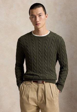 Homme portant un pull en maille torsadée vert foncé sur une chemise blanche et un pantalon plissé beige, les mains dans les poches, sur un fond neutre.