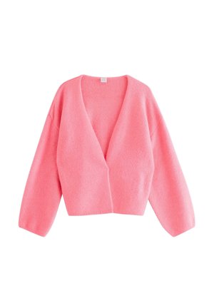 Junge Frau lächelt, trägt einen rosa Cardigan, ein weißes Spitzen-Top, weite blaue Jeans und rosa Sneaker vor einem schlichten Hintergrund.