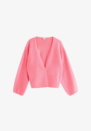 Ung kvinde smiler iført en lyserød cardigan, hvid top med blonder, viddeblå jeans og lyserøde sneakers mod en ensfarvet baggrund.