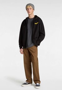 Zwarte zip-up hoodie met geel "VANS" logo, grijs T-shirt eronder, bruine broek, grijze beanie en blauwe canvas sneakers.