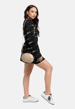 Kvinde iført en sort kjole med logomønster, hvide sneakers og en metallic guld skuldertaske, poserer med det ene ben løftet mod en ensfarvet baggrund.