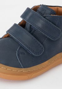 Navy blue leather shoe with two Velcro straps, rounded toe, and a tan rubber sole. Features stitching accents along the sole.