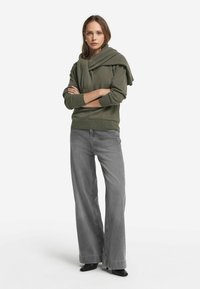 Graue weite Jeans, kombiniert mit einem dunkelgrünen Pullover, der mit einem Schal-Detail über die Schultern drapiert ist und aus weichem Strickmaterial besteht.