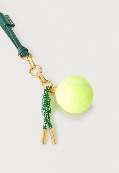 Πράσινο δερμάτινο λουρί με χρυσούς κρίκους κρατά μια μικρή λαμπερή κίτρινη μπάλα τένις και μια πράσινη φούντα με χρυσά άκρα σε λευκό φόντο.