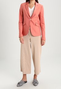 Korálový sak na míru s jedním knoflíkem a dvěma předními kapsami, v kombinaci s béžovými širokými kalhotami s plisováním a kostkovanými slip-on botami.